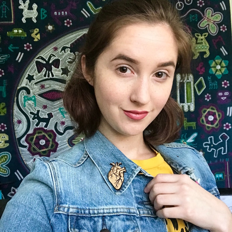 May include: A wooden anatomical heart pin, light brown in color, is pinned to a denim jacket. The pin is detailed with the veins and chambers of the heart. The person is wearing a yellow shirt underneath the jacket. A tapestry with various shapes and colors is in the background.