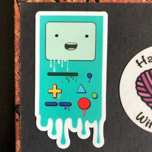 May include: A blue and green cartoon character sticker with a face and buttons. The character is dripping with a white liquid.