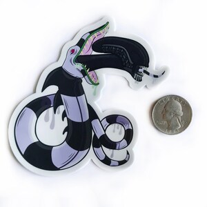 May include: A sticker of a purple and black striped creature with a large mouth and a green tongue. The creature is eating a black alien with a white skull. The sticker is about the size of a US quarter.