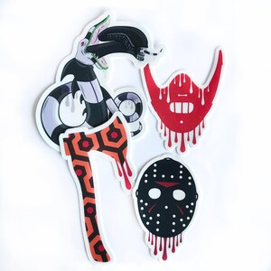 May include: Four horror movie themed stickers. A red and black axe with a pattern, a red dripping beard, a black and white hockey mask with red dripping blood, and a black and green snake with a mouth open.