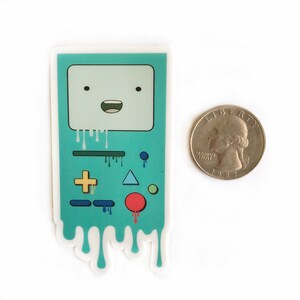 May include: A teal blue sticker of a video game console with a smiling face and dripping paint. The console has a plus sign, a triangle, a circle, and a line on the front. The sticker is about the size of a US quarter.