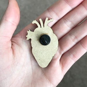 May include: A wooden anatomical heart pin with a black pin back. The pin is light brown with a detailed outline of the heart's chambers and vessels. The pin is held in a person's hand.