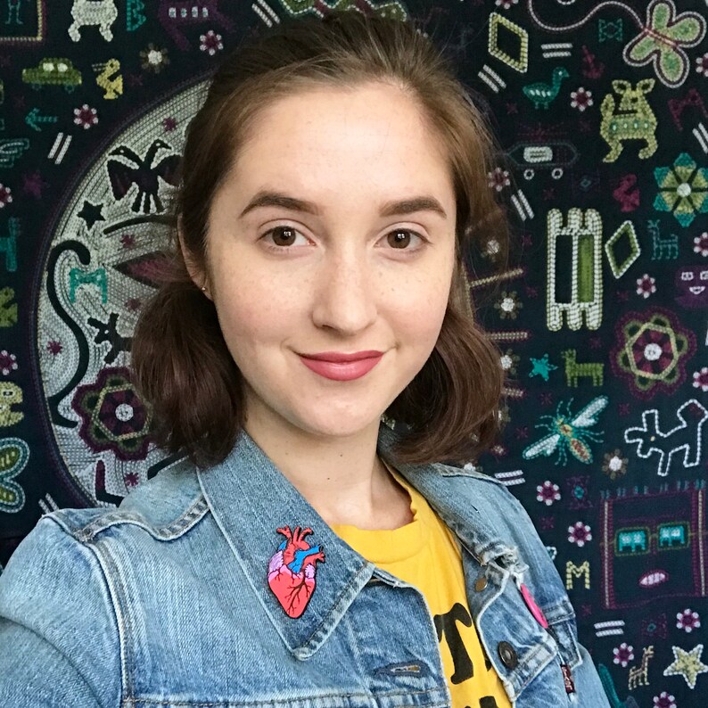 May include: A woman wearing a denim jacket and a yellow t-shirt with a graphic. A colorful anatomical heart pin is attached to the jacket. The background features a tapestry with various patterns and designs.
