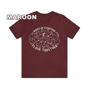 K&ouml;nnte beinhalten: Ein maroonfarbenes T-Shirt mit einer wei&szlig;en Grafik von H&uuml;hnern und dem Text "Birds of a feather flock together".
