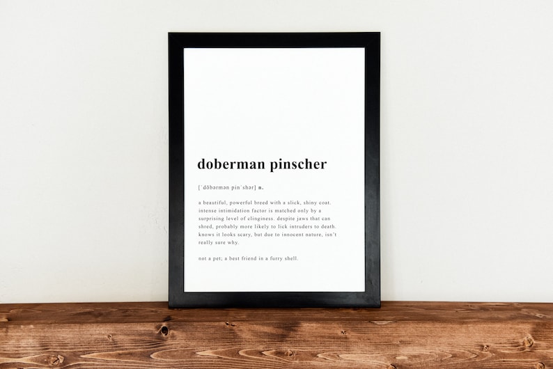 May include: A black and white printable definition of the word "Doberman Pinscher" in a black frame. The definition describes the breed as a "beautiful, powerful breed with a slick, shiny coat" and notes that they are "probably more likely to lick intruders to death".