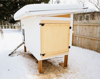May include: A white chicken coop with a wooden door and a metal ramp. The coop is on wooden legs and is sitting on a snowy ground.