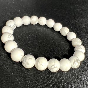 May include: White beaded bracelet with gray veining. The bracelet is made of round, polished beads. The beads are strung together to form a circular shape. The bracelet is on a black background.