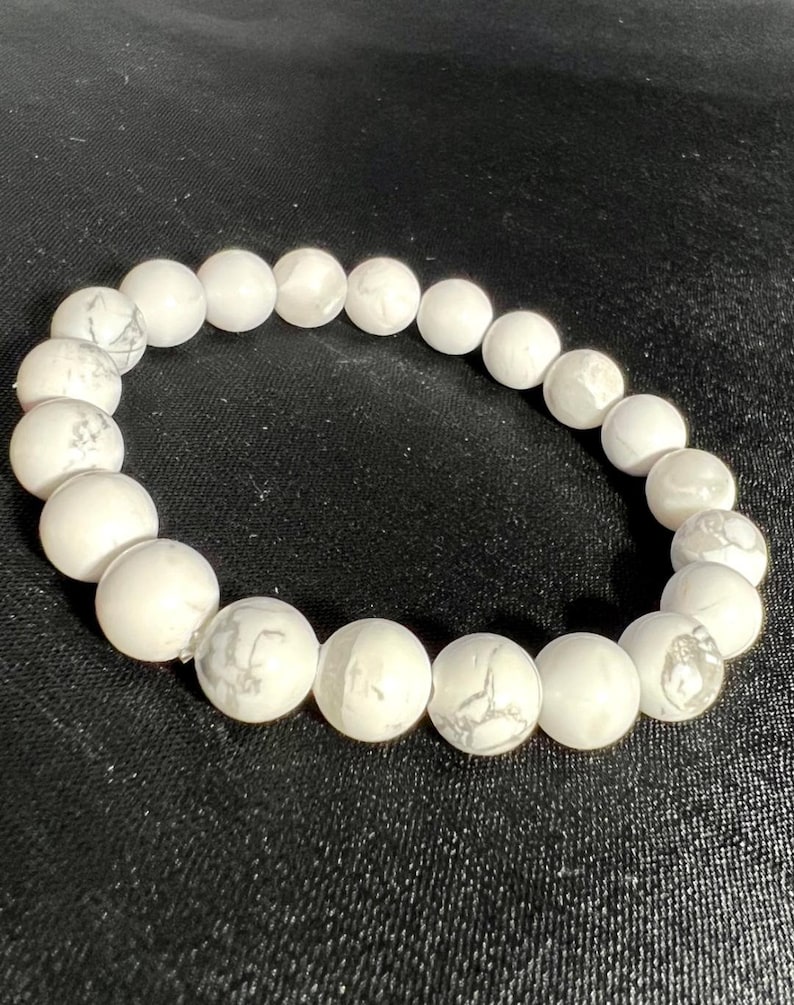 May include: White beaded bracelet with gray veining. The bracelet is made of round, polished beads. The beads are strung together to form a circular shape. The bracelet is on a black background.