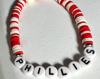 May include: A red and white striped bracelet with a baseball charm and the word "PHILLIES" spelled out in white beads.