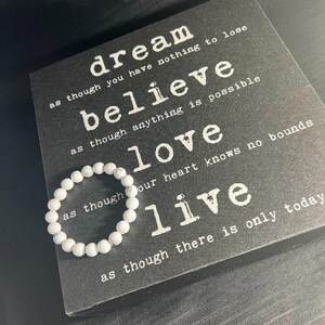 May include: A white beaded bracelet rests on a black decorative sign with white text. The sign features the words "dream, believe, love, live" with inspirational phrases. The bracelet is made of small, round, white beads.
