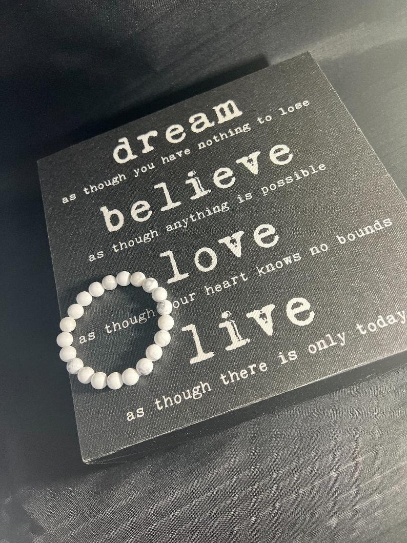 May include: A white beaded bracelet rests on a black decorative sign with white text. The sign features the words "dream, believe, love, live" with inspirational phrases. The bracelet is made of small, round, white beads.
