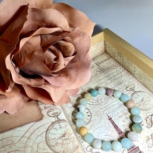 Puede incluir: Un brazalete de piedras preciosas azul claro y verde con un acento de piedra preciosa marr&oacute;n claro. El brazalete est&aacute; sobre un libro vintage con un dise&ntilde;o floral y una esfera de reloj.