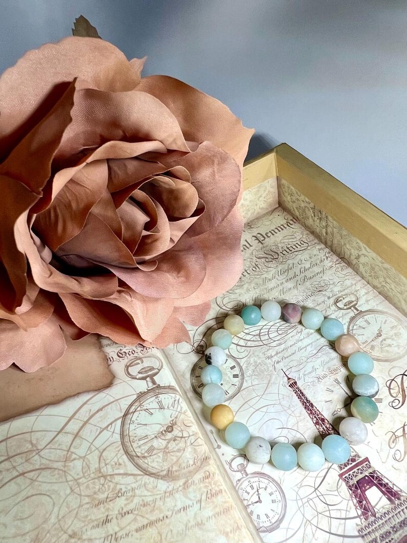 Puede incluir: Un brazalete de piedras preciosas azul claro y verde con un acento de piedra preciosa marr&oacute;n claro. El brazalete est&aacute; sobre un libro vintage con un dise&ntilde;o floral y una esfera de reloj.