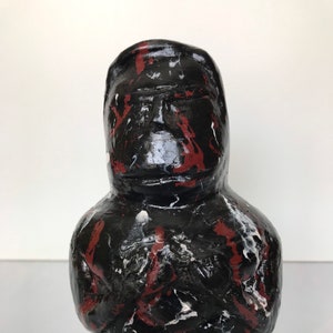 Może przedstawiać: Czarna ceramiczna rzeźba przedstawiająca osobę z czerwonymi i białymi plamami farby. Rzeźba ma zaokrąglony, ludzki kształt z twarzą i ramionami.
