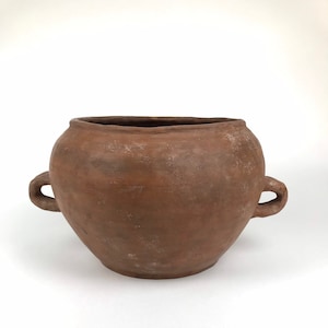 Può includere: Un vaso rustico in terracotta marrone con un'ampia apertura e due manici ad anello. Il vaso ha un corpo arrotondato e una superficie strutturata, che suggerisce un aspetto invecchiato. Ideale per l'arredamento della casa o il giardinaggio.