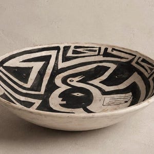 Op de afbeelding: Een witte keramische kom met een zwart geometrisch en vogelontwerp. De ovale kom heeft een brede rand. Het zwarte ontwerp bedekt de binnenkant en creëert een opvallend contrast met de witte achtergrond. De kom is waarschijnlijk met de hand beschilderd.