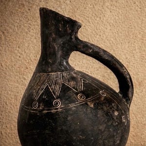 Op de afbeelding: Een donkere, antieke keramische kruik met een handvat en een tuit. De zwarte kruik heeft een decoratief wit geëtst ontwerp rond de body en hals. De kruik heeft een afgeronde basis en een licht schuine tuit.