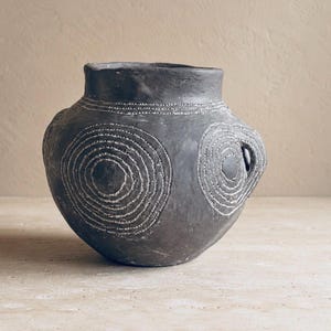 Op de afbeelding: Een donker antracietkleurige keramische pot met een ronde vorm en een brede opening. Het buitenoppervlak is voorzien van met de hand uitgesneden concentrische cirkelpatronen die met wit pigment zijn geaccentueerd voor een textuurcontrast.