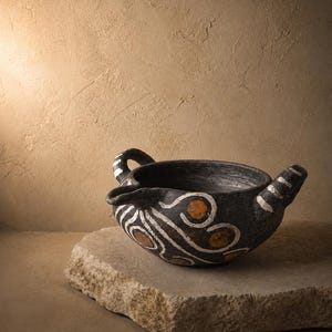 Puede incluir: Un cuenco de cerámica negra con dos asas y un pico, decorado con patrones de remolinos blancos y naranjas. El cuenco está sobre una piedra tosca, con un fondo neutro.