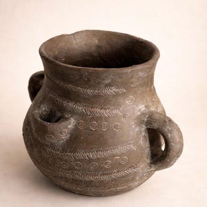Può includere: Un vaso di ceramica marrone in stile antico, con due manici e un beccuccio. È decorato con motivi incisi a forma di piume e cerchi. Il recipiente ha un'ampia apertura e una consistenza leggermente ruvida.