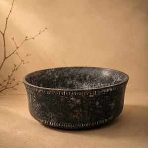 Op de afbeelding: Een donkere, getextureerde kom met een brede rand en een licht uitlopende vorm. De kom heeft een verweerde, gevlekte uitstraling met tinten zwart, bruin en grijs. De rand en de basis hebben een decoratief, stikselachtig patroon.