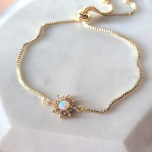 May include: A gold bracelet featuring a delicate chain and a central sunburst charm. The charm has a central opal-like stone. The bracelet is displayed on a white, marble-effect surface.