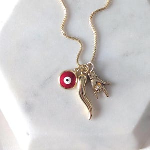 May include: A gold-tone chain necklace featuring three charms: a circular red and white evil eye pendant, a smooth gold-tone curved horn-shaped charm, and a gold-tone hand charm displaying a gesture with the index and pinky fingers extended.