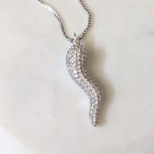 May include: A silver-toned necklace featuring a curved, horn-shaped pendant entirely encrusted with small, clear, sparkling stones. The pendant hangs from a delicate, thin silver-toned box chain, creating a cohesive and reflective metallic appearance.
