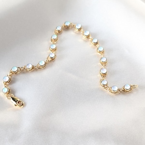 May include: A delicate gold bracelet featuring a series of round, opalescent stones. The bracelet has a decorative, openwork design around each stone and a secure clasp. The jewelry is displayed on a white, silky background.