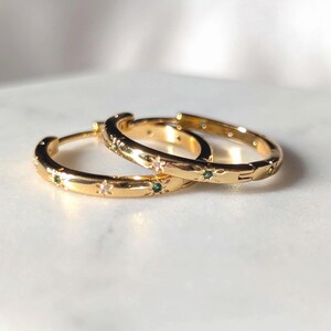 May include: A pair of gold hoop earrings featuring a polished finish with embedded small star-shaped accents. Each star is set with either a clear white stone or a small green stone, creating an alternating pattern along the circular band.