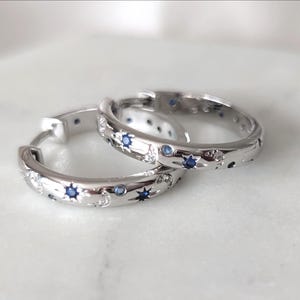 May include: A pair of silver hoop earrings with a celestial design. Each earring features small blue and clear gemstones set in a pattern of stars and dots. The earrings are displayed on a white marble surface.