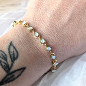 May include: A gold-colored bracelet with oval-shaped, opalescent stones. The bracelet is linked with a delicate, textured design. The stones are set in a gold-colored bezel. The bracelet is worn on a wrist with a black tattoo.
