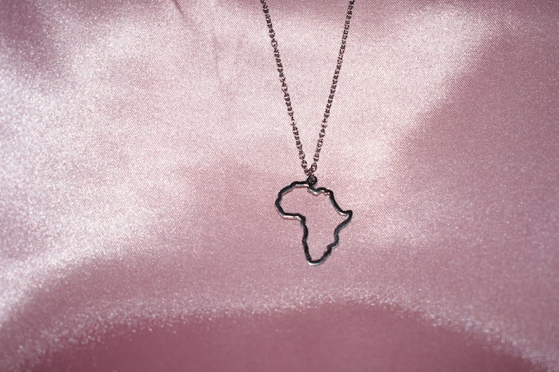 May include: A silver chain necklace with an outline of the continent of Africa as the pendant. The necklace is displayed on a pink satin background.
