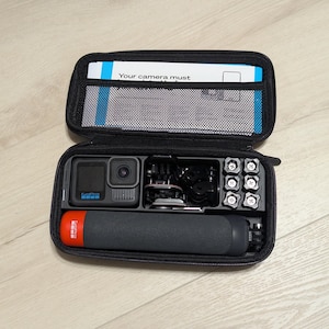 Op de afbeelding: Een zwarte draagtas met een GoPro-camera, een selfie-stick en diverse accessoires. De tas heeft een ritssluiting en een gaasvak aan de binnenkant. De tekst "Your camera must" is zichtbaar aan de binnenkant van de tas.