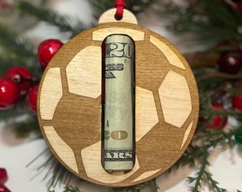 Könnte beinhalten: Ein Holz-Fußball-Anhänger mit einem Zwanzig-Euro-Schein darin. Der Anhänger hängt an einem roten Band und ist von grünen Kiefernästen umgeben.