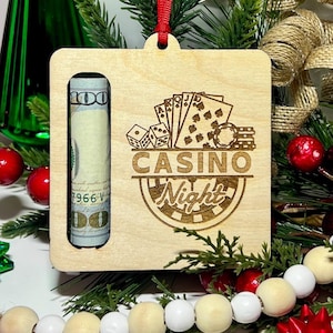May include: A wooden Christmas ornament with a slot to hold a rolled-up dollar bill. The ornament is engraved with the words "Casino Night" and a roulette wheel design. The ornament is hanging from a red ribbon.