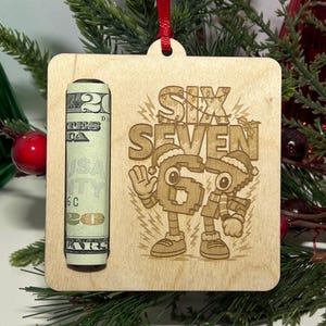 May include: A wooden Christmas ornament with a rolled-up twenty-dollar bill and the words "Six Seven" with cartoon characters. The ornament is square with a red ribbon for hanging. The background includes Christmas decorations.