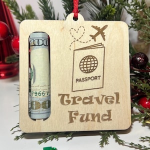 May include: Wooden Christmas ornament with a slot for cash, a passport and airplane design, and the text "Travel Fund".