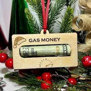 May include: A wooden Christmas ornament shaped like a gas gauge with a rolled up dollar bill inside. The ornament is labeled "GAS MONEY" and has a Santa hat icon.