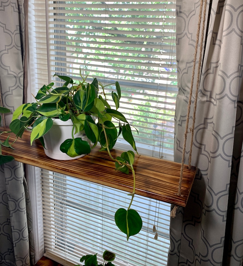 May include: A hanging wooden shelf with a potted plant in front of a window. The shelf is suspended by rope and has a natural wood finish. The plant has green and yellow leaves. The window has blinds and is framed by patterned curtains.