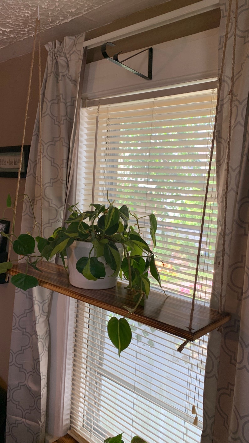 May include: A wooden hanging shelf with a white potted plant, suspended by rope in front of a window. The plant has green and yellow leaves. The shelf is stained a dark brown color.