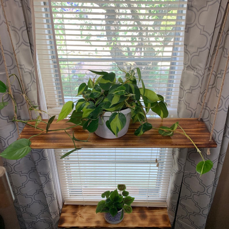 May include: A two-tiered wooden hanging shelf with a natural finish. The top shelf holds a white pot with a green and yellow plant. The bottom shelf has a small plant in a patterned pot. The shelves are suspended by rope.