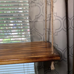 May include: A wooden shelf with a rich brown stain, suspended by thick, natural-fiber ropes. The shelf is rectangular and appears to be made of multiple wooden planks. The ropes are attached to the shelf's corners. The background includes a window with blinds and a patterned curtain.