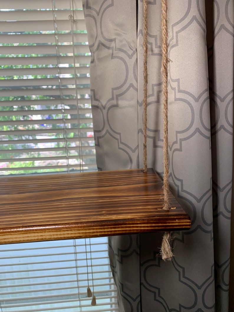 May include: A wooden shelf with a rich brown stain, suspended by thick, natural-fiber ropes. The shelf is rectangular and appears to be made of multiple wooden planks. The ropes are attached to the shelf's corners. The background includes a window with blinds and a patterned curtain.