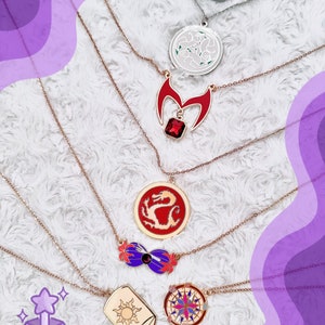 May include: A set of seven gold-tone necklaces with various charms, including a dragon, a compass, a sun, a crown, a magic wand, and a flower. The necklaces are displayed on a white fluffy surface.