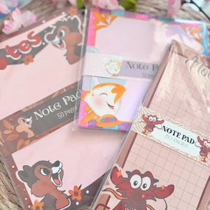 Può includere: Quattro blocchi note diversi con vari personaggi Disney. I blocchi note sono rosa, marroni e beige con accenti marroni, neri e rossi. I blocchi note presentano personaggi dei film Disney: I Rescuers, Mulan e Bambi. Ogni blocco note ha il testo "Note Pad 50 Pages".