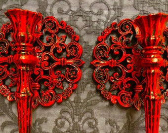 May include: Two ornate, red metal wall sconces with black accents. Each sconce features a candle holder and a decorative, floral-patterned backplate. The sconces are mounted on a textured, gray background.