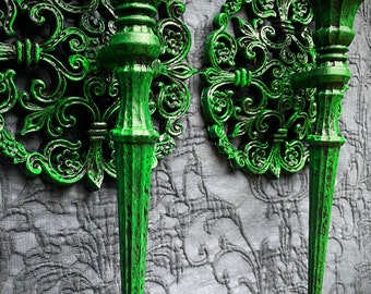 May include: A pair of ornate green metal wall sconces with a scrolled design. The sconces are in the shape of a torch and have a decorative base.