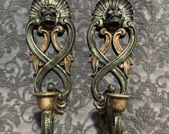 May include: Pair of ornate wall sconces with a skull design. The sconces are made of metal with a distressed finish in shades of silver, gold, and bronze. Each sconce features a skull at the top, intricate scrollwork, and a candle holder.