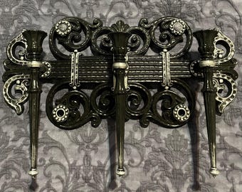 May include: Ornate black wall sconce with three candle holders. The sconce features scrolling designs and silver accents. Each candle holder has a long, tapered shape with a white tip. The overall design is symmetrical and decorative.